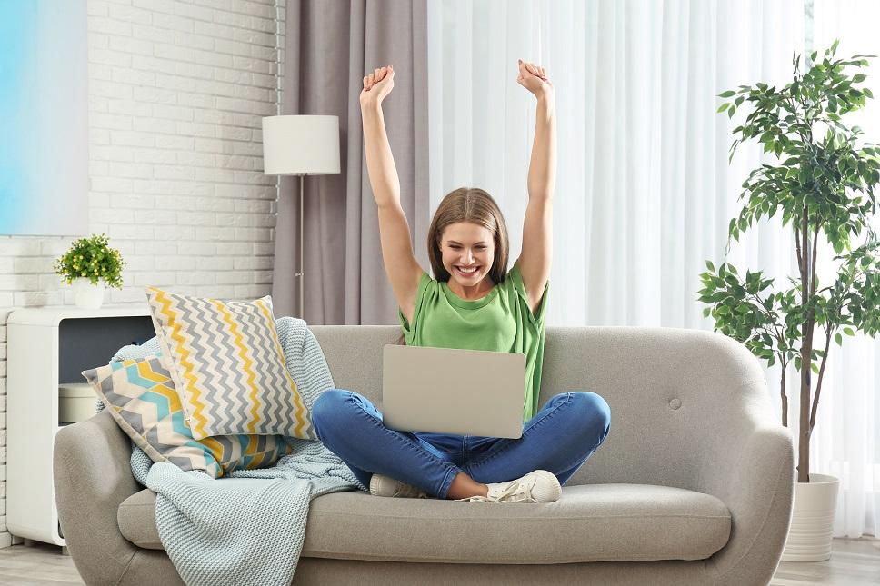 Emotional young woman with laptop celebrating victory on sofa at home